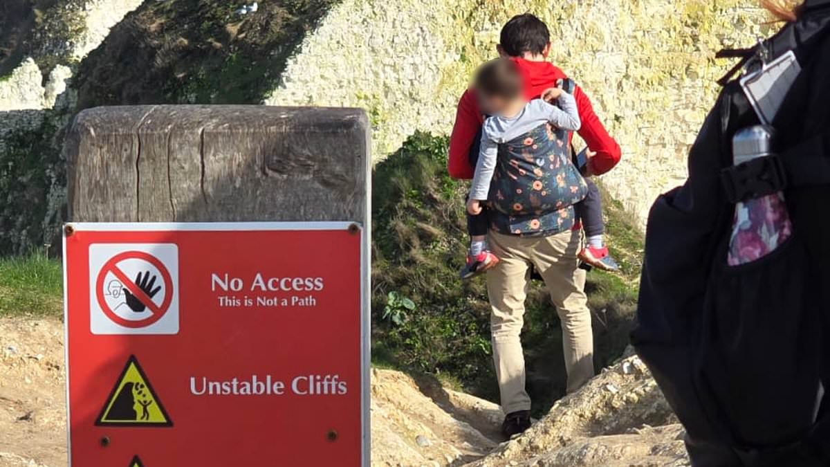 Foolhardy father carrying young child in backpack risks their lives by ignoring warning sign to get clifftop view